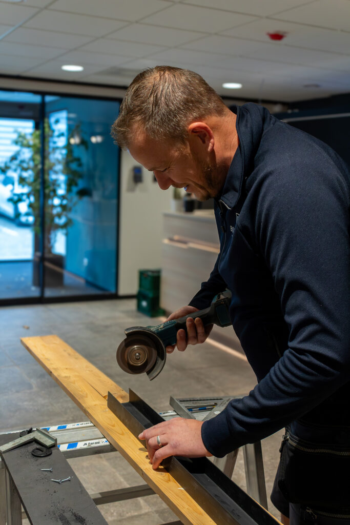 Een man slijpt zorgvuldig een metalen profiel op een werkbank in een moderne, goed verlichte werkruimte.
