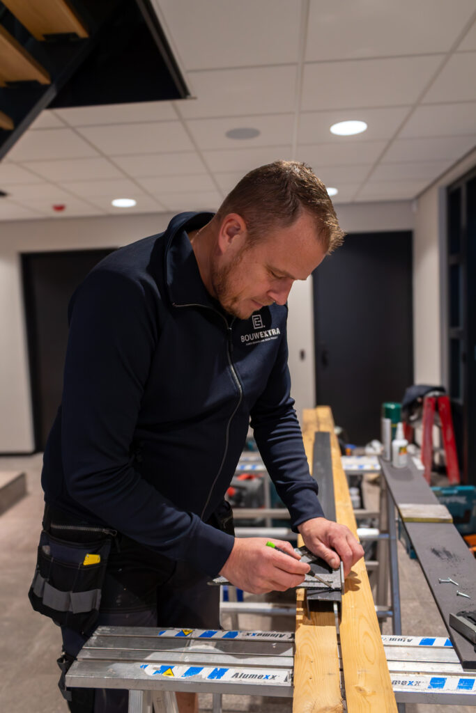 Een medewerker van Bouwextra meet en markeert zorgvuldig een houten balk met een stalen hoekliniaal in een werkplaats.