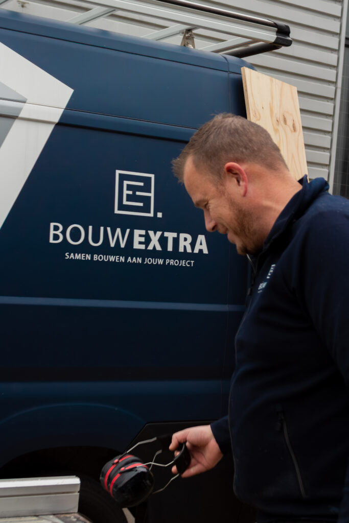 Een medewerker van Bouwextra staat buiten bij een bedrijfsbus en houdt een gehoorbeschermer in zijn hand.