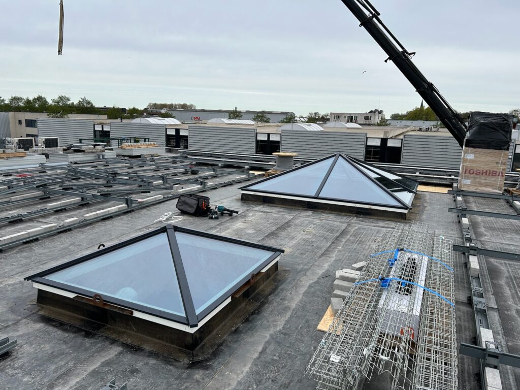 Een plat dak met metalen draagconstructies en twee piramidevormige lichtkoepels in aanbouw op een bedrijventerrein.